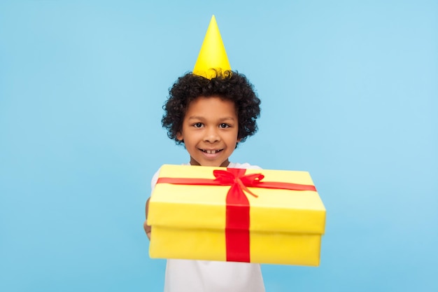 Generoso y adorable niñito con un divertido cono de fiesta en la cabeza sonriendo felizmente y dando regalos a la cámara, saludando con cumpleaños, compartiendo una gran caja de regalos para ti. foto de estudio interior fondo azul