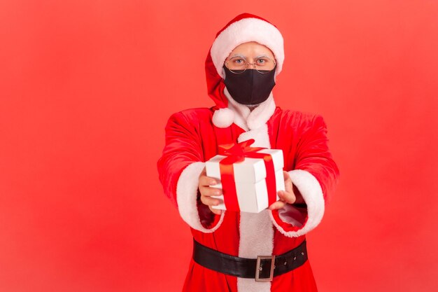 Generoso anciano santa claus con anteojos y máscara protectora en la cara sosteniendo y mostrando una caja de regalo con lazo rojo haciéndole un regalo Foto de estudio interior aislada sobre fondo rojo