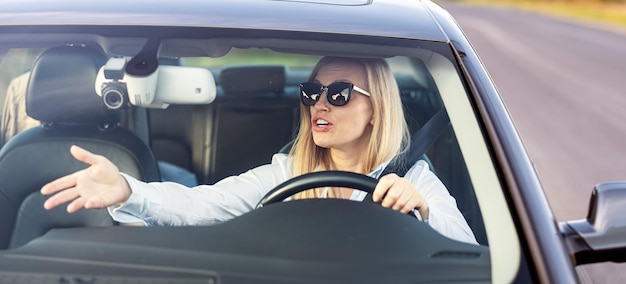 Genervte reife Frau in Business-Outfit und Sonnenbrille, die mit der Hand gestikuliert und jemanden anschreit, während sie ihr Auto fährt Probleme mit dem Straßen- und Verkehrskonzept