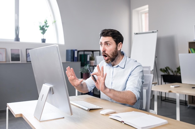 Genervter emotionaler nervöser Büroangestellter eines jungen Mannes, der über einen Videoanruf am Computer spricht, der in weißer Kleidung an einem Tisch im Büro sitzt