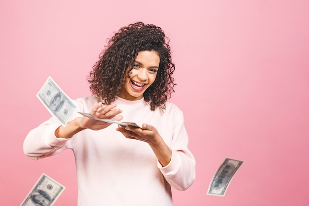 ¡Genial chica rica! ¡Ganador de dinero! Hermosa mujer afroamericana sorprendida en casual sosteniendo dinero en gafas de sol aisladas sobre fondo rosa.