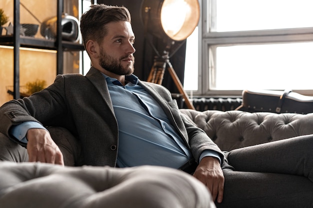 Genial y guapo. Apuesto joven en traje completo sentado en el sofá mientras pasa tiempo en el interior.