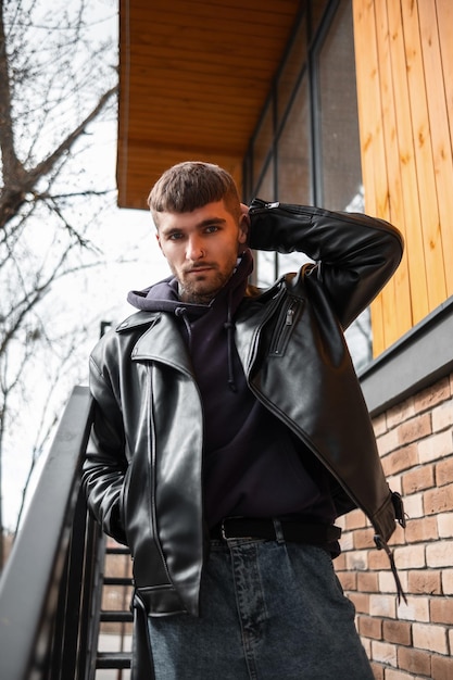 Genial hipster brutal con una chaqueta negra de cuero de moda y una sudadera con capucha en una escalera de metal fuera de un edificio en la calle
