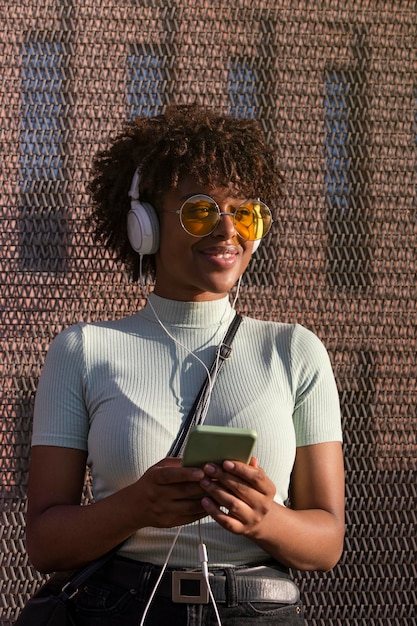 Genial joven mujer latinoamericana con cabello afro y gafas sonriendo escuchando música en un teléfono inteligente con auriculares