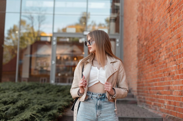 Genial modelo de mujer hipster de moda con gafas de sol de moda en una elegante chaqueta de cuero y top con gafas de sol vintage camina cerca de un edificio de ladrillos de un centro comercial