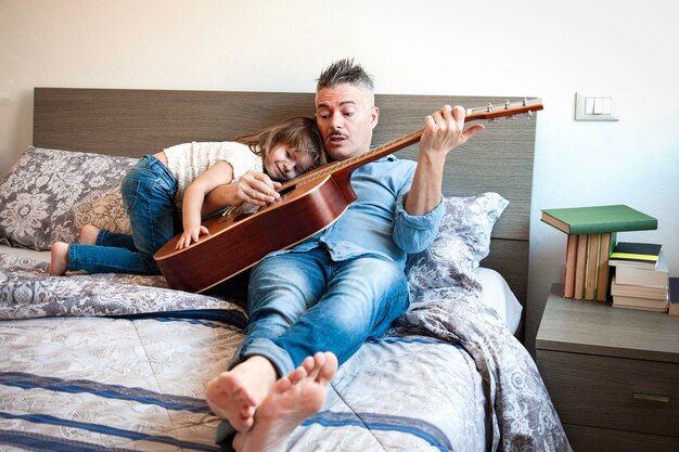 Genial papá tocando la guitarra acústica y enseñando una canción mientras su hija lo escucha Papá e hija están sentados en la cama tocando la guitarra Concepto de enseñanza y paternidad Día del padre