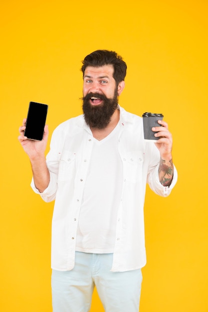 Genial rutina matutina feliz hombre barbudo sosteniendo el teléfono mientras bebe café brutal hipster bebiendo bebida para llevar de un tipo de taza con barba y bigote mostrando teléfono móvil