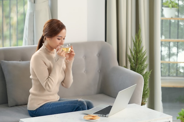 Genieße das Wochenende. Asiatisches Mädchen, das sich entspannt, Kaffee trinkt und Laptop benutzt, zu Hause auf der Couch sitzt sitting