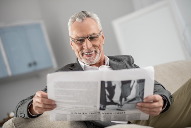 Geniesse dein Leben. Hübsche männliche Person, die zu Hause ist und Lächeln auf seinem Gesicht hält, während Artikel liest