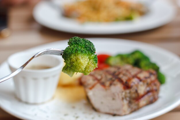 Genießen Sie, Bissen des Brokkolis mit Gabel und Schweinefleisch saftigem Steakhintergrund zu essen