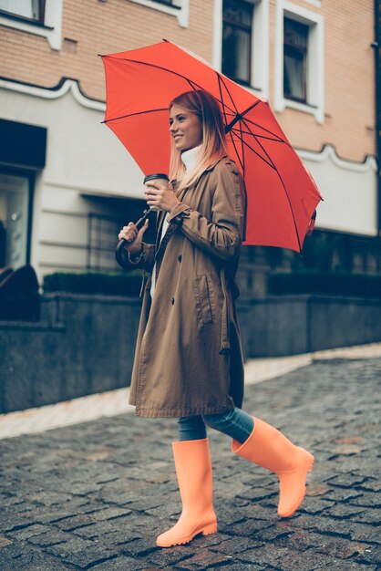 Genießen Sie den Herbst in der Stadt. Volle Länge der attraktiven jungen lächelnden Frau in Gummistiefeln mit Regenschirm und Kaffeetasse, während sie die Straße entlang geht