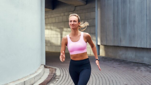 Genießen Sie die junge lächelnde Frau in bequemer Sportkleidung, die draußen auf der Straße läuft