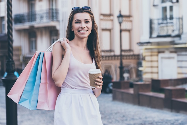 Genießen Sie frischen Kaffee nach dem Shopping. Schöne junge Frau, die Einkaufstüten trägt und eine Kaffeetasse hält, während sie im Freien steht