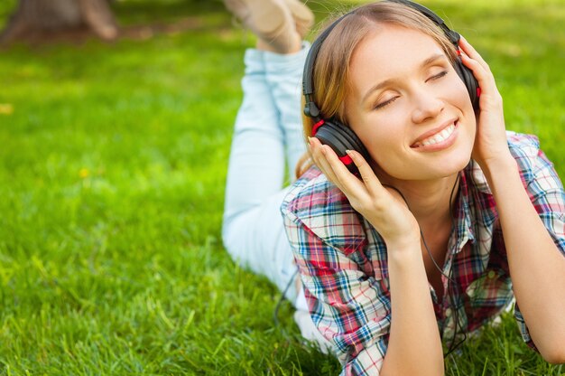 Genießen Sie Ihre Lieblingsmusik in der Natur. Schöne junge Frau mit Kopfhörern, die Musik hört und lächelt, während sie auf dem grünen Gras liegt