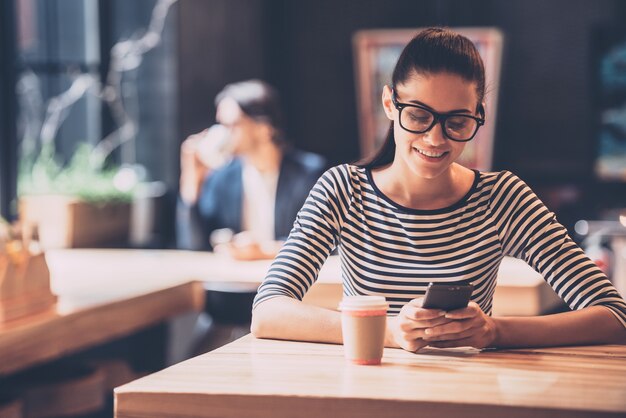 Genießen Sie kostenloses WLAN. Fröhliche junge Frau, die ein Smartphone hält und es mit einem Lächeln betrachtet, während sie im Café sitzt und im Hintergrund Kaffee trinkt