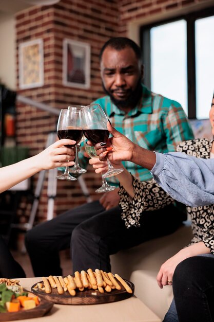 Gente alegre brindando copas de vino mientras celebra su cumpleaños en casa. Diverso grupo de amigos cercanos en la fiesta del vino tintineando vasos mientras disfrutan de la diversión juntos en la sala de estar.