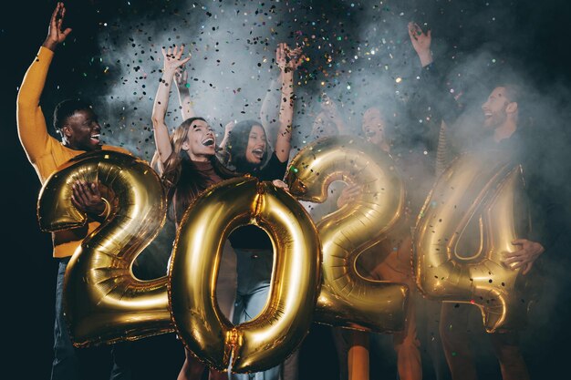 Gente alegre con números de 2024 y lanzando confeti mientras celebran el Año Nuevo en un club nocturno