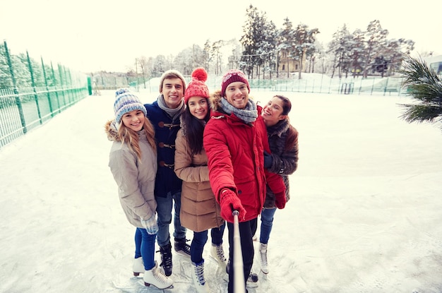 gente, amistad, tecnología y concepto de ocio - amigos felices tomando fotos con un selfie de smartphone en una pista de patinaje sobre hielo al aire libre