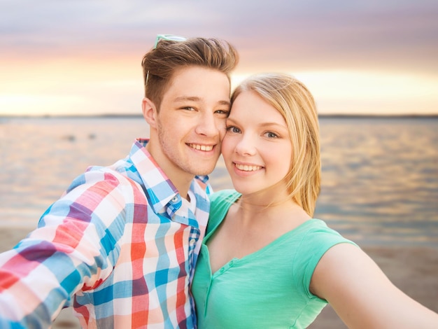 gente, amor, mar, tecnología y concepto de vacaciones de verano - pareja feliz tomando selfie con smartphone o cámara sobre el fondo de la playa al atardecer