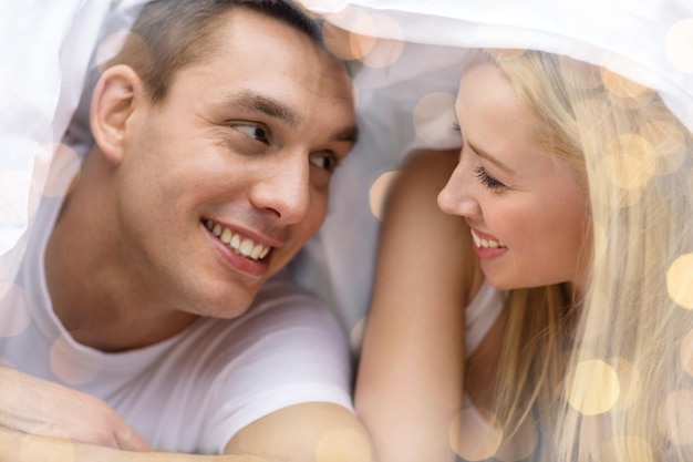 gente, amor, relaciones y concepto de hora de acostarse - pareja feliz bajo una manta en la cama sobre fondo de luces