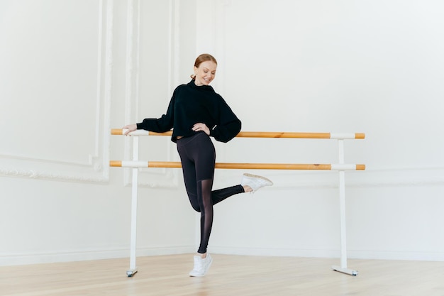 Gente bailando concepto de flexibilidad de coreografía Delgada mujer pelirroja encantada con traje negro levanta una pierna cerca de la barra de ballet en prácticas de estudio de danza vacías antes de la actuación