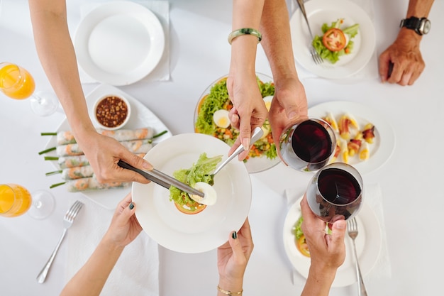 Gente bebiendo vino y comiendo sabrosa ensalada de verduras y rollitos de primavera en la cena, vista desde la parte superior