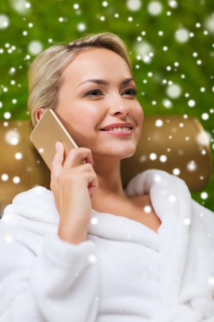 gente, belleza, estilo de vida, tecnología y concepto de relajación - hermosa mujer joven en bata de baño blanca con teléfono inteligente llamando y hablando en el spa con efecto de nieve