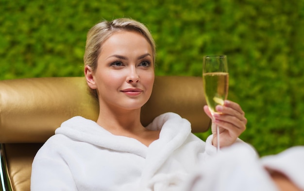 gente, belleza, estilo de vida, vacaciones y concepto de relajación - hermosa mujer joven en bata de baño blanca tumbada en la tumbona y bebiendo champán en el spa