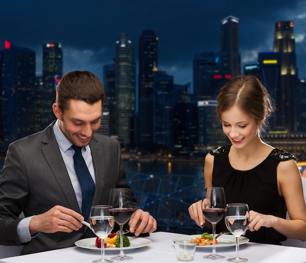 gente, celebración, concepto romántico y de vacaciones - pareja sonriente comiendo plato principal con vino tinto en el restaurante durante la noche de fondo de la ciudad