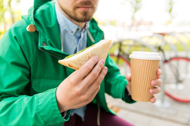 gente, comida basura, comida y estilo de vida - cerca de un joven con una taza de café y un sándwich comiendo y bebiendo en la calle de la ciudad