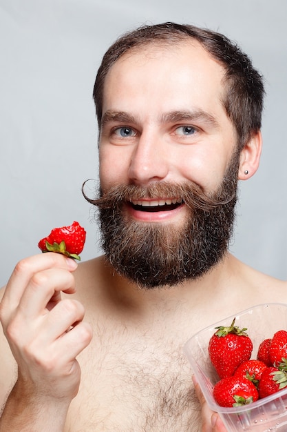 Gente, comida, belleza, estilo de vida, concepto de día de San Valentín - retrato de primer plano joven sosteniendo una fresa y sonriendo. Retrato de guapo barbudo y bigote. sobre un fondo gris