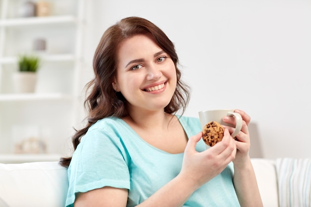 Foto gente, comida chatarra, alimentación poco saludable y concepto de ocio - mujer joven feliz de talla grande con taza de té con galleta en casa