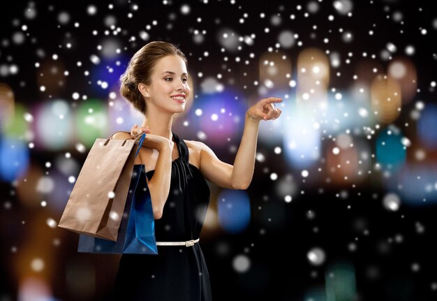 gente, compras, venta, concepto de Navidad y vacaciones - mujer sonriente vestida con bolsas de compras sobre luces nocturnas y fondo de nieve