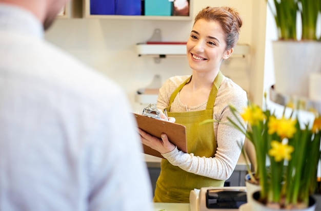 Foto gente, compras, venta, floristería y concepto de consumismo - mujer florista sonriente feliz con portapapeles y hombre o cliente haciendo pedidos en la floristería