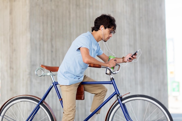 gente, comunicación, tecnología y estilo de vida - hombre hipster con teléfono inteligente, auriculares y taza termo en bicicleta fija escuchando música en la calle de la ciudad