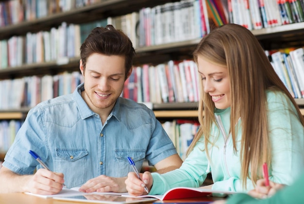 gente, conocimiento, educación y concepto escolar - grupo de estudiantes felices escribiendo en cuadernos en la biblioteca