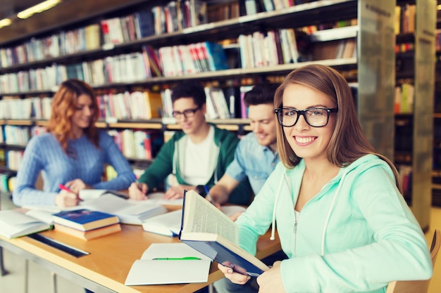 Foto gente, conocimiento, educación, literatura y concepto escolar - mujer joven feliz con anteojos leyendo un libro y preparándose para los exámenes sobre un grupo de estudiantes en la biblioteca