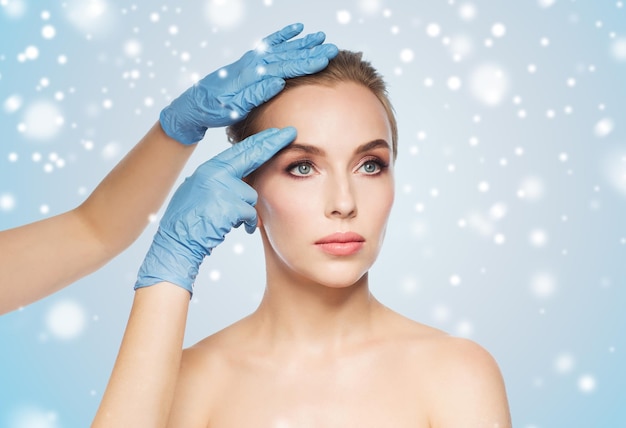 gente, cosmetología, cirugía plástica y concepto de belleza - manos de cirujano o esteticista tocando la cara de la mujer sobre fondo azul y nieve