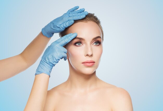 gente, cosmetología, cirugía plástica y concepto de belleza - manos de cirujano o esteticista tocando la cara de la mujer sobre fondo azul