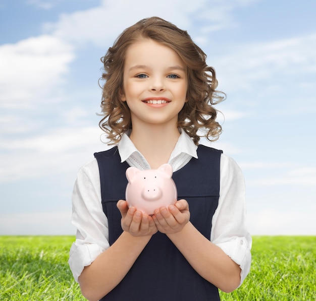 gente, dinero, finanzas y concepto de ahorro - niña feliz sosteniendo una alcancía en las palmas sobre el cielo azul y el fondo de la hierba