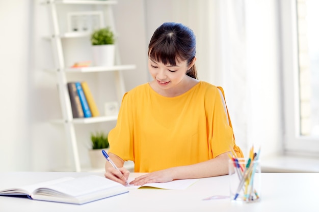 gente, educación, escuela secundaria y concepto de aprendizaje - feliz joven estudiante asiática con libro y libreta escribiendo en casa