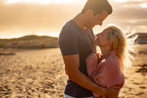 Gente enamorada y puesta de sol dorada al aire libre - pareja de hombre y mujer se abrazan y miran con la playa de fondo - concepto de protección y vida juntos para siempre