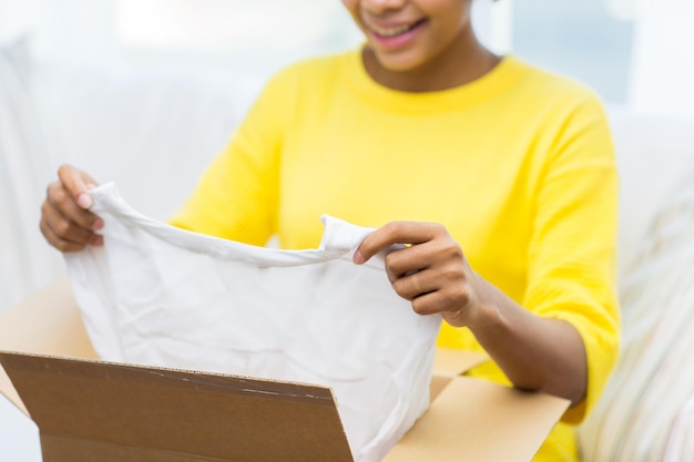 gente, entrega, comercio, envío y concepto de servicio postal - cerca de una joven afroamericana feliz sacando ropa de una caja de cartón o un paquete en casa