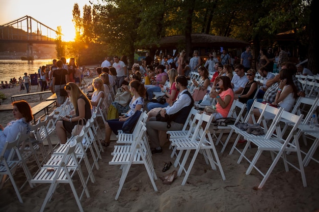 La gente está viendo un concierto de música clásica. Mujeres y hombres jóvenes y adultos borrosos y otras personas que miran escuchando un concierto de una banda clásica en un ambiente al aire libre con luz diurna.