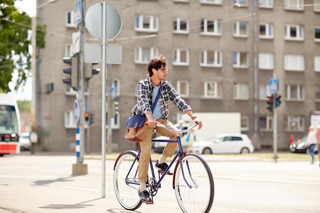 gente, estilo, ocio y estilo de vida - joven hipster con bolsa de hombro y auriculares montando una bicicleta de engranajes fijos en la calle de la ciudad