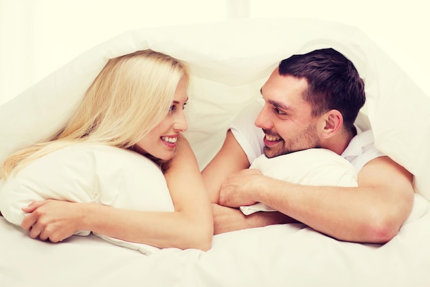gente, familia, hora de acostarse y concepto de felicidad - pareja feliz acostada en la cama cubierta con una manta sobre la cabeza y hablando en casa