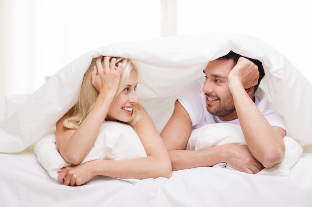 gente, familia, hora de acostarse y concepto de felicidad - pareja feliz acostada en la cama cubierta con una manta sobre la cabeza y hablando en casa