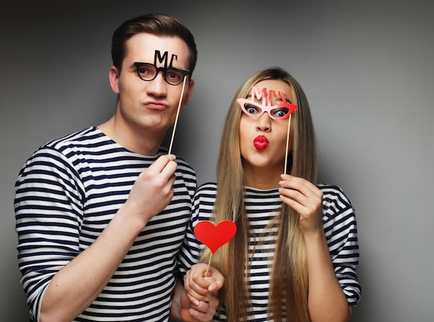 Gente fiesta amor y ocio concepto encantadora pareja sosteniendo gafas de fiesta y sombrero en palos sobre fondo gris