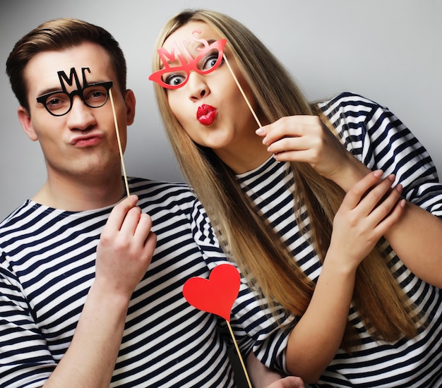 Gente fiesta amor y ocio concepto encantadora pareja sosteniendo gafas de fiesta y sombrero en palos sobre fondo gris