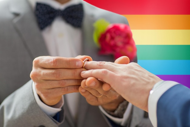 gente, homosexualidad, matrimonio entre personas del mismo sexo y concepto de amor - cerca de las manos de una pareja gay masculina feliz poniendo el anillo de bodas sobre el fondo de la bandera del arco iris